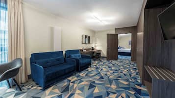 A hotel room with a blue sofa set, a desk with a chair, a flat-screen TV, and a geometric patterned carpet.