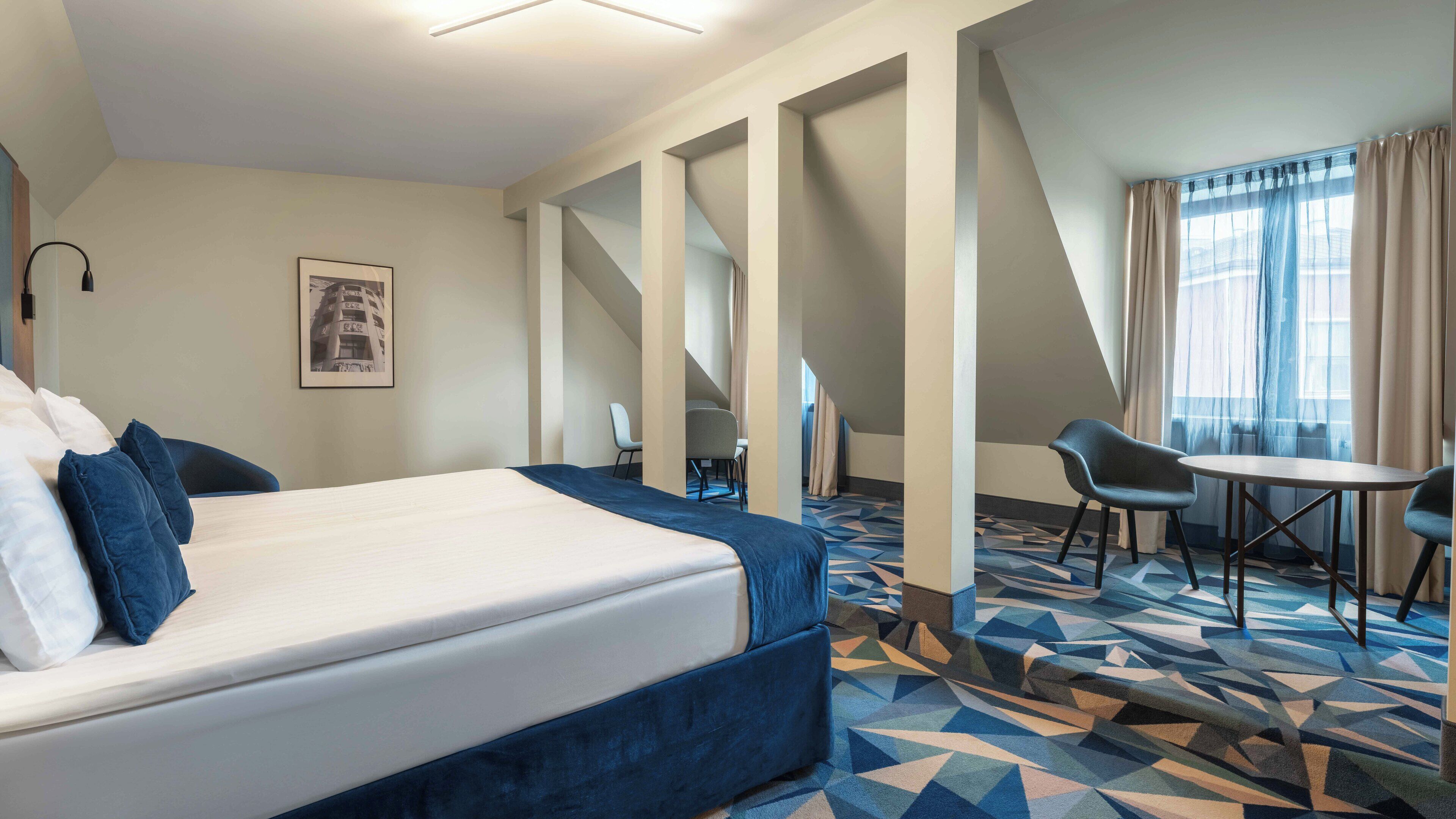 A hotel room with a bed, a desk, two chairs, a patterned carpet, and a large window with curtains.