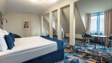 A hotel room with a bed, a desk, two chairs, a patterned carpet, and a large window with curtains.