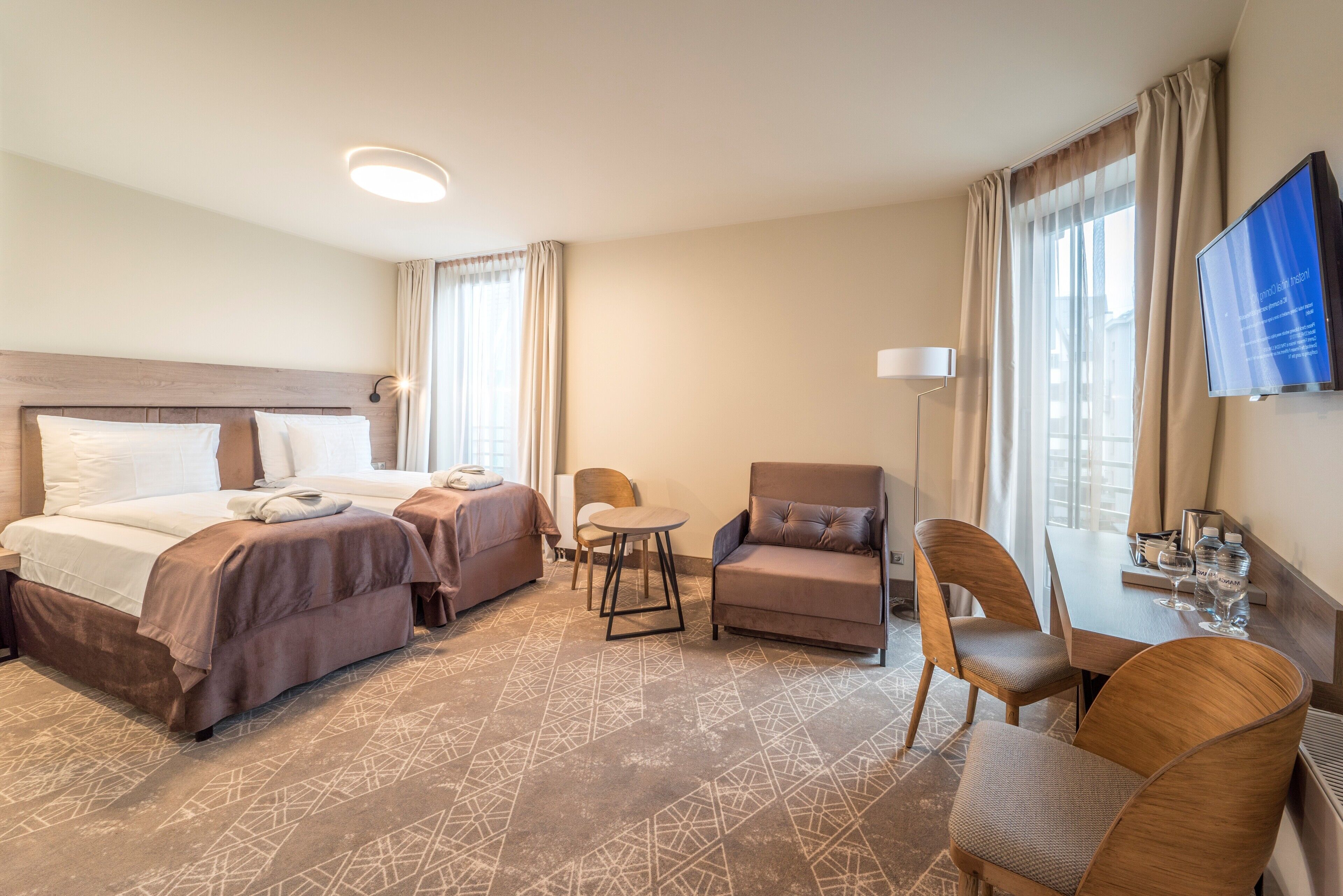 A hotel room with two beds, a sofa, a chair, a small table, a TV, and a window with curtains.