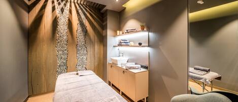 Een moderne spa-ruimte met een massagetafel, een houten kastje met handdoeken en een spiegel die de ruimte weerspiegelt.