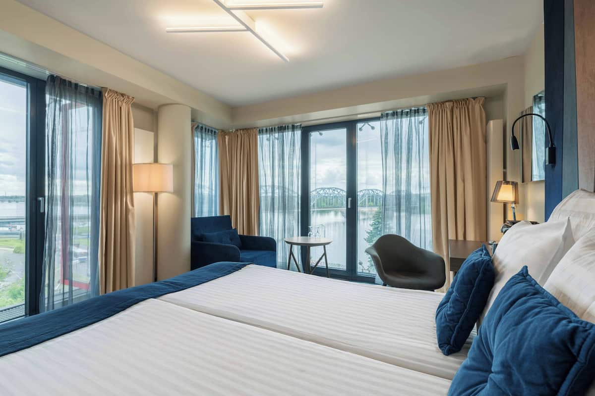 A hotel room with a large bed, two chairs, a small table, and large windows offering a view of a bridge and water.