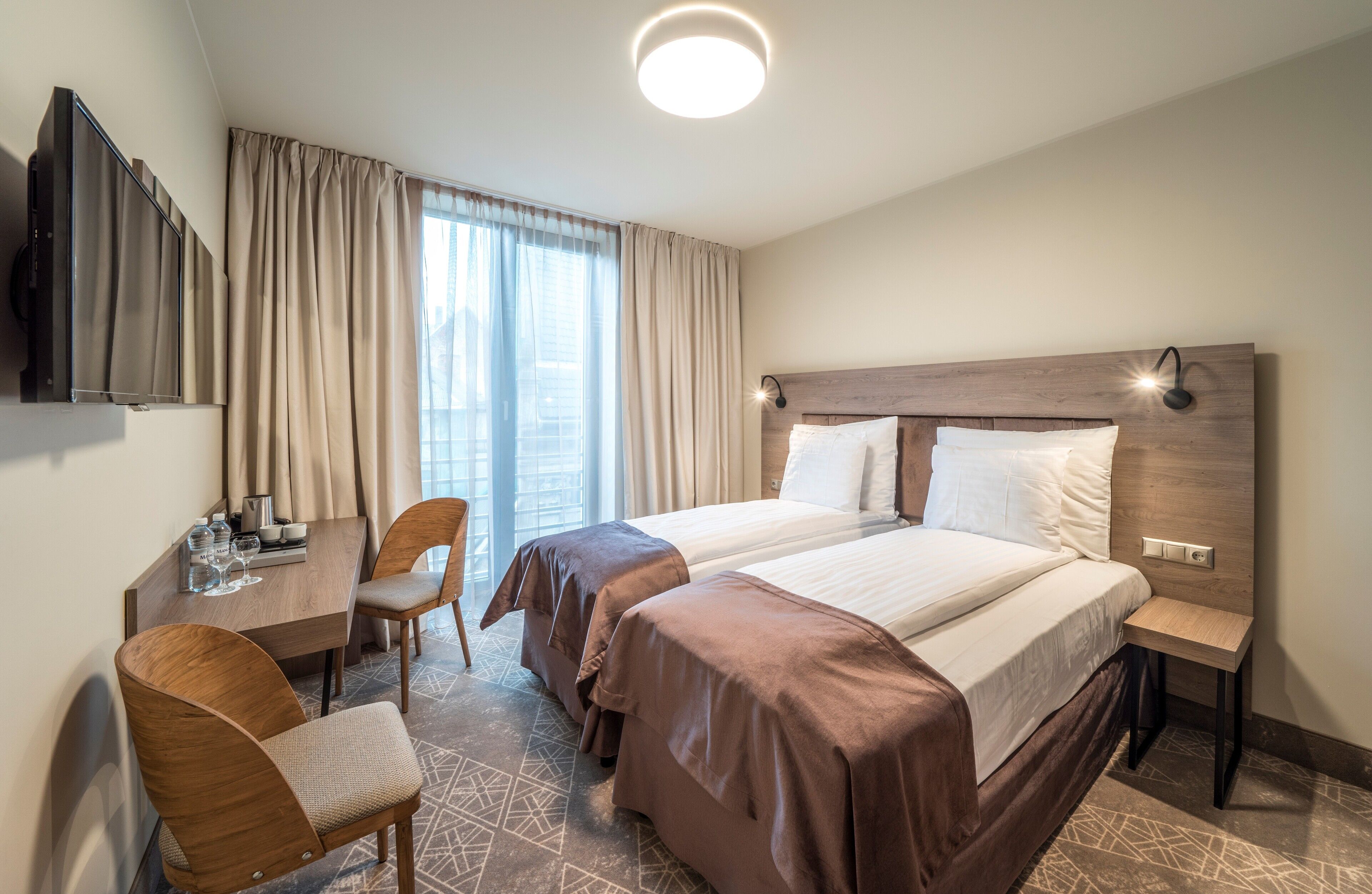 A hotel room with two beds, a desk with a chair, a TV, and a large window with curtains.
