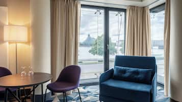A hotel room with a large window, a blue armchair, a purple chair, a small table with two wine glasses, and a lamp.