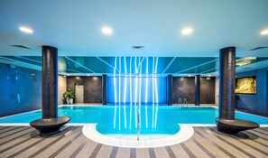 An indoor swimming pool with a modern design, featuring a central water feature, two large columns, and a patterned floor.