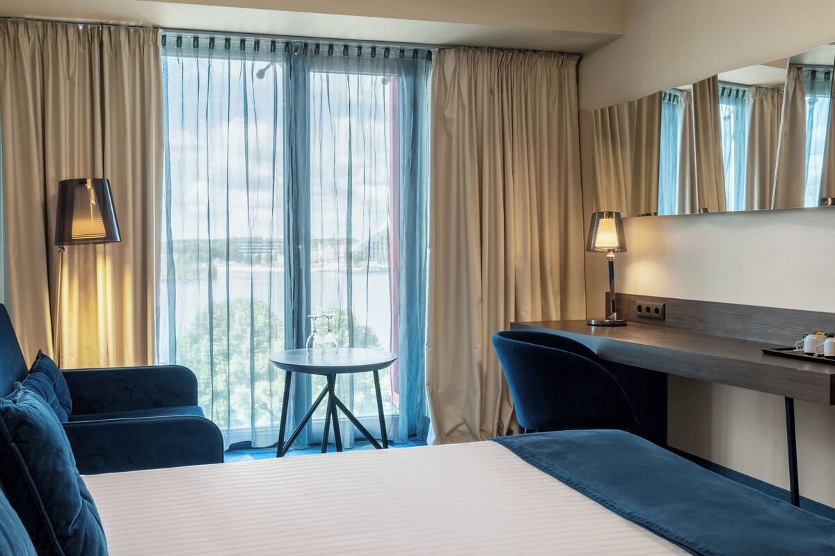 A hotel room with a bed, a desk, a chair, a table, and a view of a body of water through sheer curtains.
