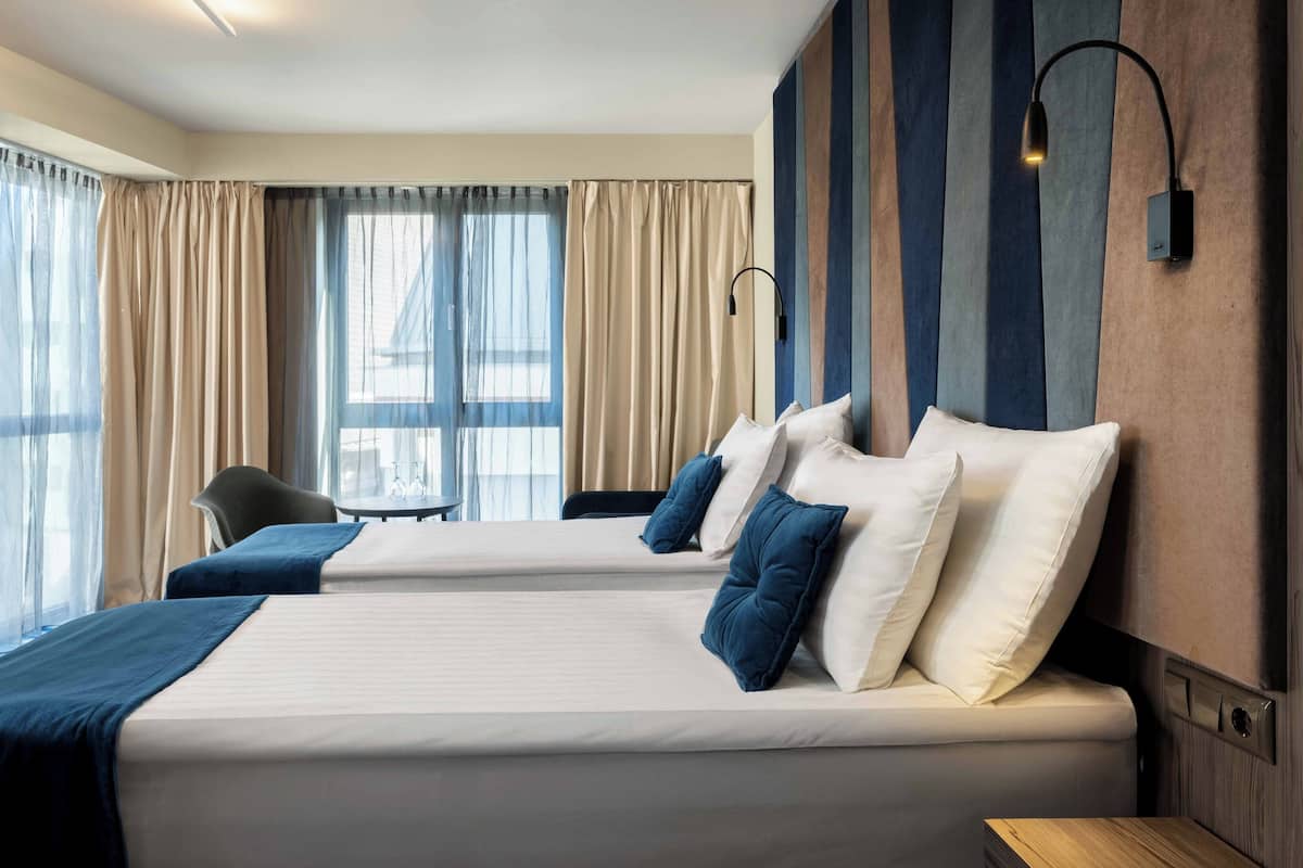 A hotel room with two beds, a chair, a table, and a large window with curtains.