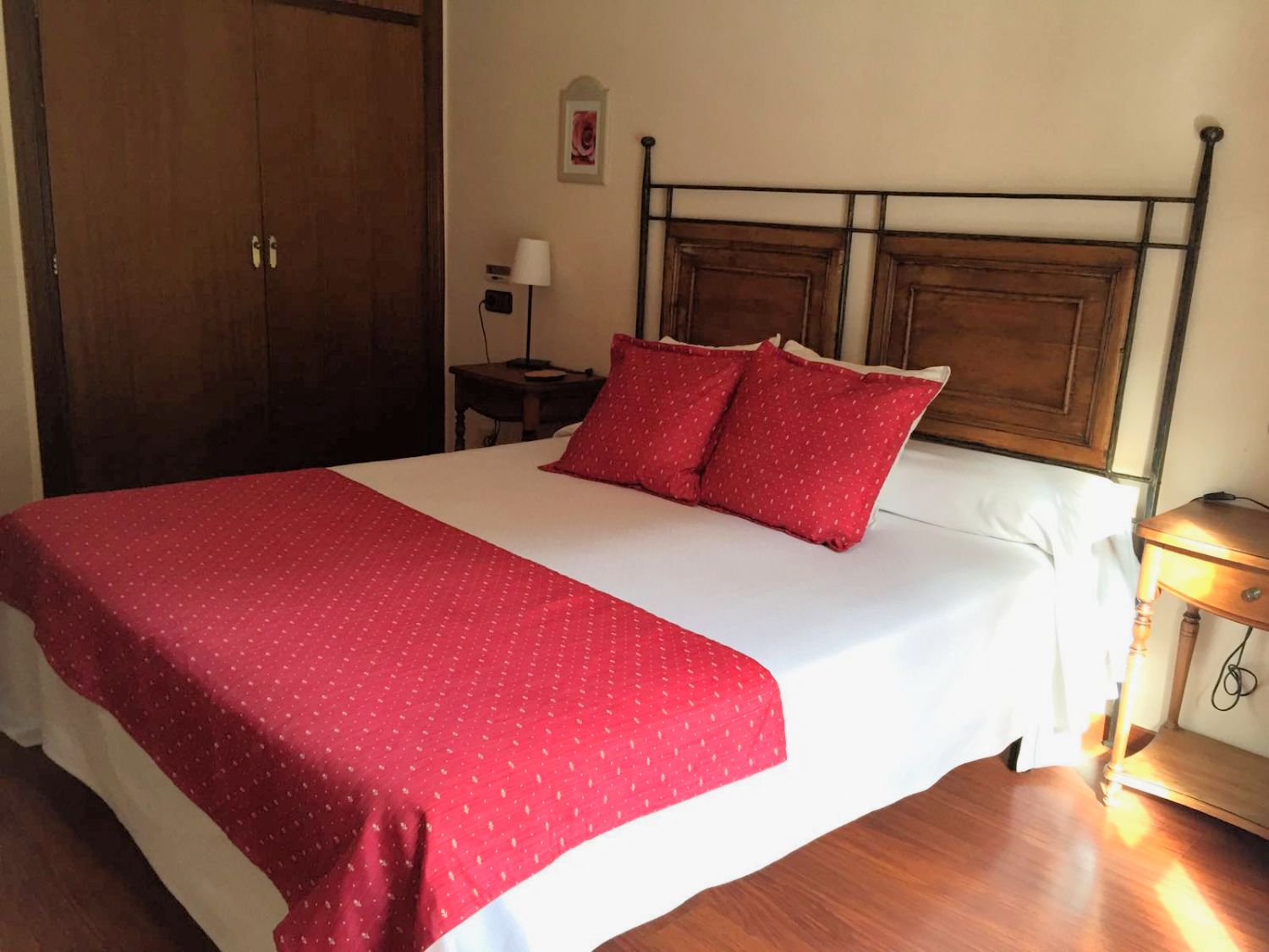 A neatly made bed with a red blanket and pillows, a wooden headboard, a bedside table with a lamp, and a wooden wardrobe.