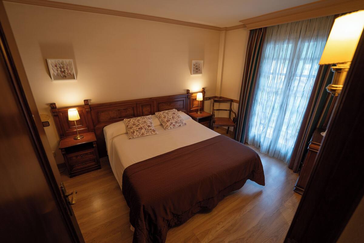 Quarto de hotel com uma cama grande, duas mesas de cabeceira com abajures, cabeceira de madeira e uma cadeira perto da janela.