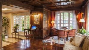 A spacious room with wooden flooring, a large window with curtains, a desk with a laptop, a dining table with chairs, and a sofa.