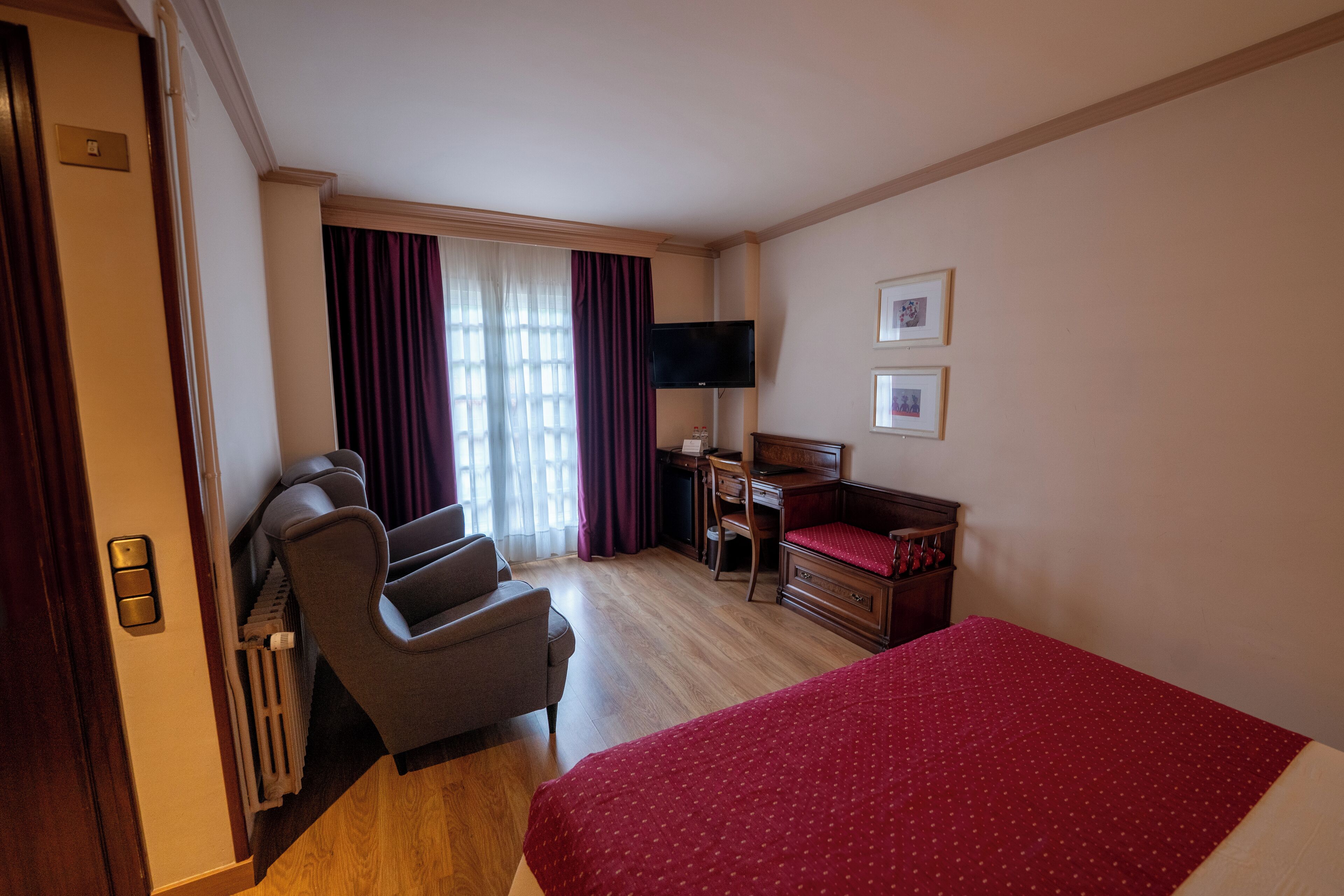 A hotel room with a bed, a desk with a chair, a TV, and a window with curtains.