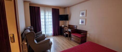 A hotel room with a bed, a desk with a chair, a TV, and a window with curtains.