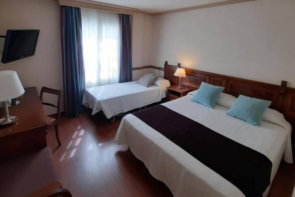 Quarto de hotel com duas camas de solteiro, uma escrivaninha com cadeira, televisão e janela com cortinas.