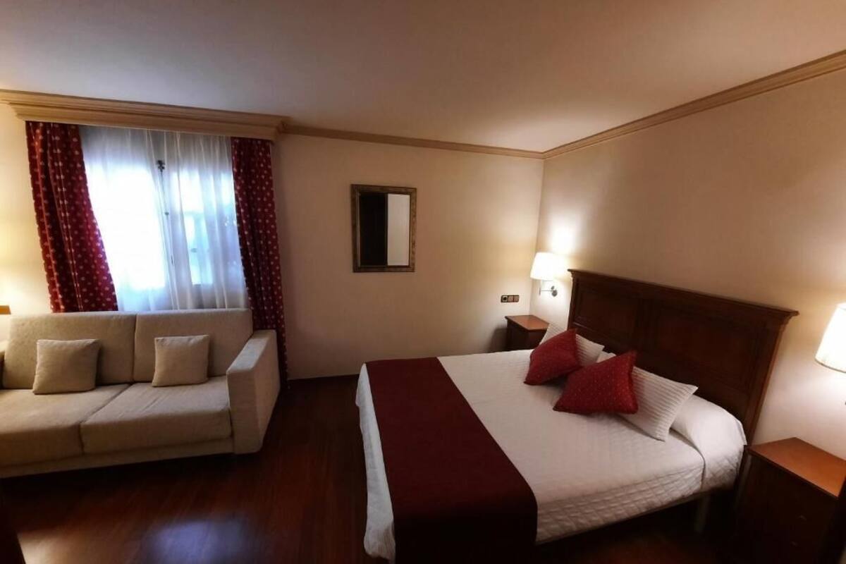 Quarto de hotel com cama, sofá, mesa de cabeceira, luminária e janela com cortinas.