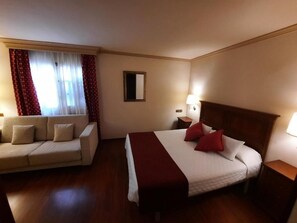 A hotel room with a bed, a sofa, a nightstand, a lamp, and a window with curtains. - Hotel Edelweiss Camprodon (Camprodon)