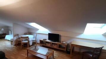 A cozy loft-style living space with a wooden floor, a sofa, a dining table with chairs, a TV, and a desk.