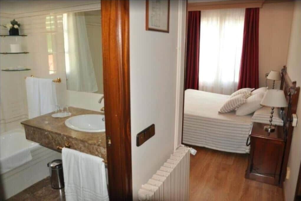 Habitación de hotel con baño, cama, mesita de noche y ventana con cortinas.