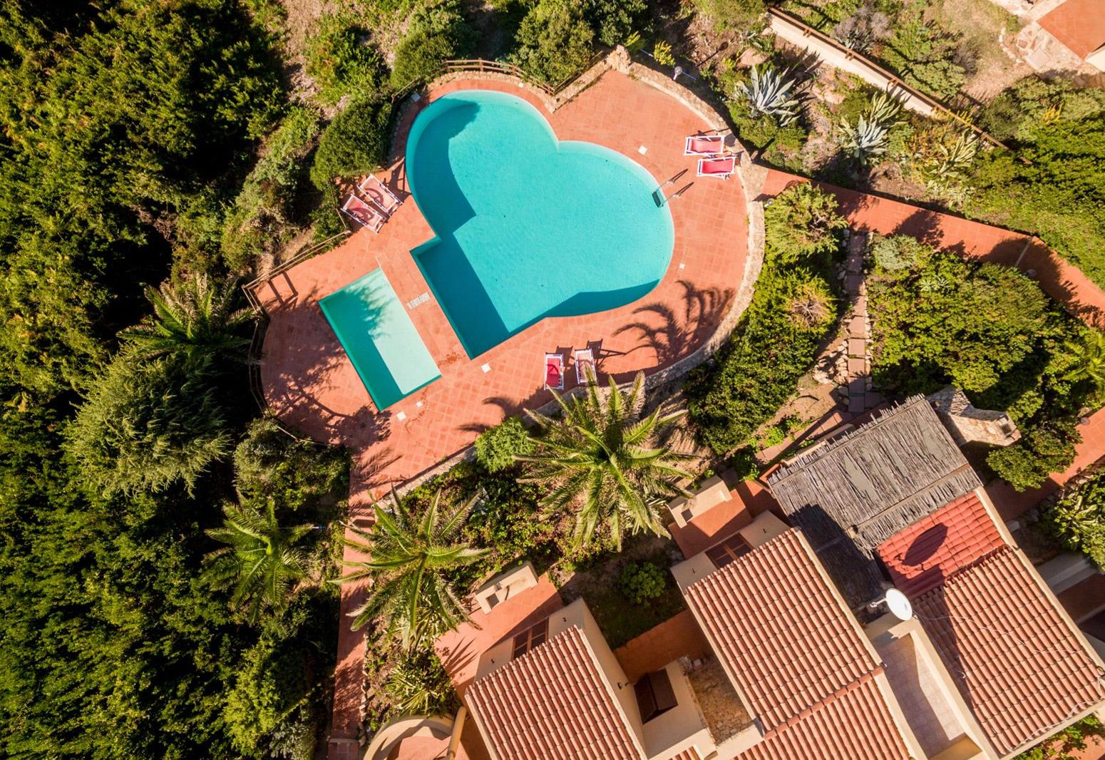 An aerial view of a resort with a large swimming pool, palm trees, and red-roofed buildings.