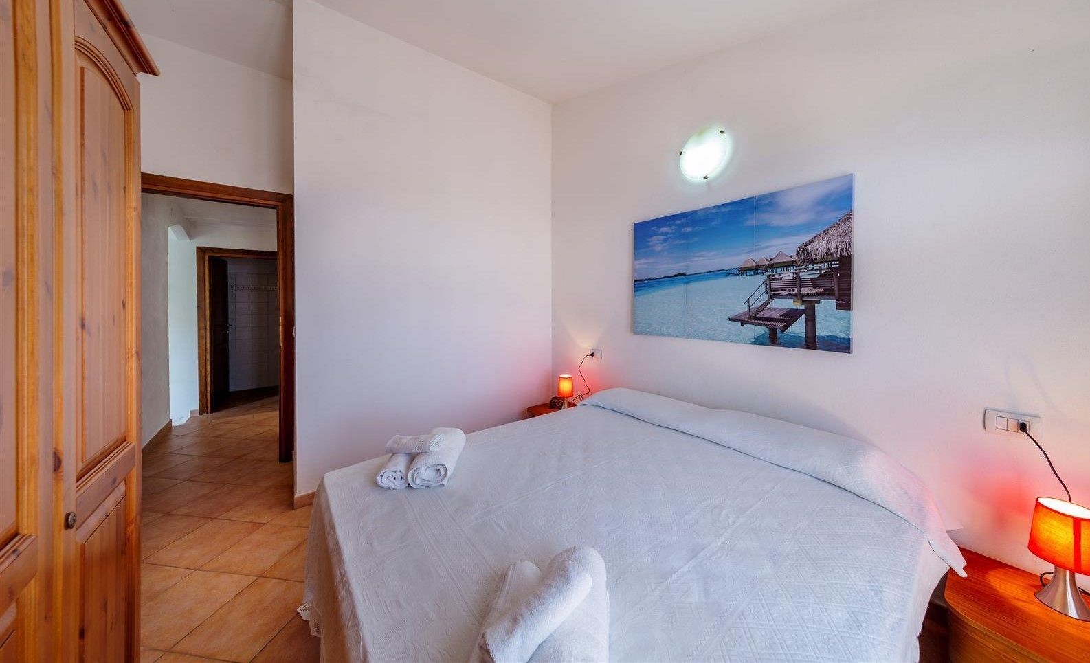 Una habitación de hotel con una cama, un cuadro de una playa, un armario de madera y una mesita de noche con una lámpara.