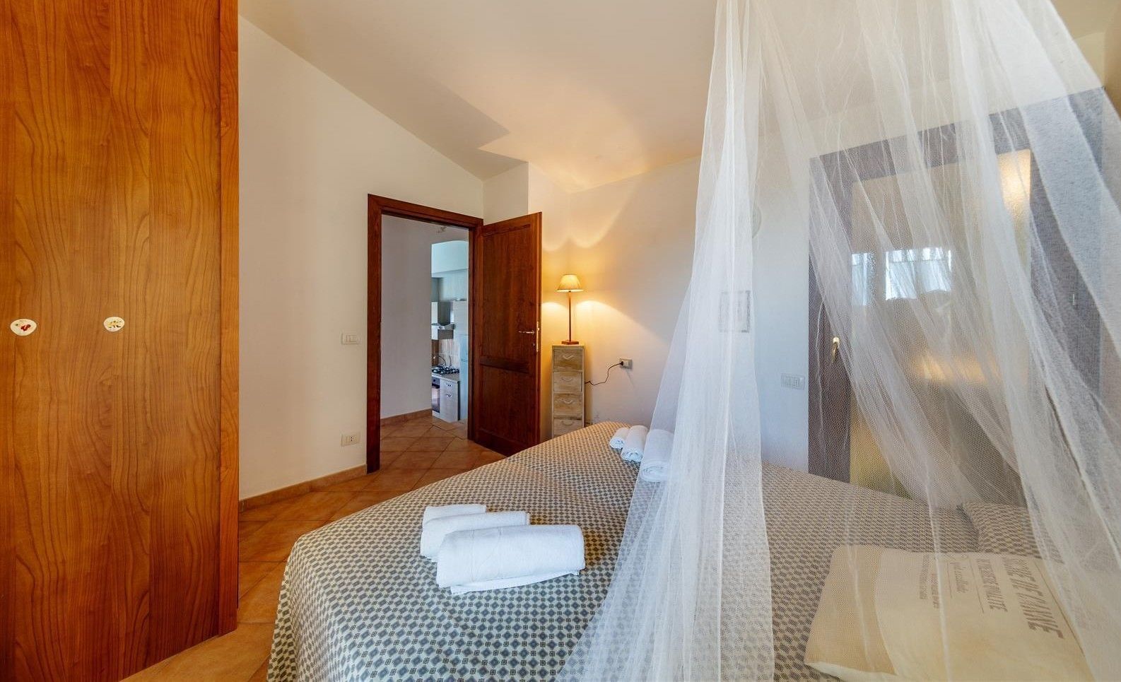 Ein Schlafzimmer mit einem Bett, einer Holztür, einem Spiegel, einer Nachttischlampe und einem Fenster mit durchscheinendem Vorhang.