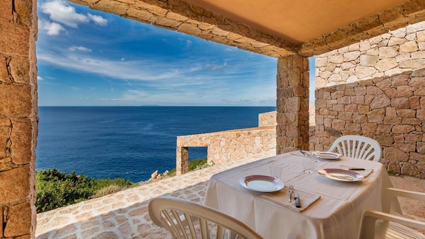 A stone-walled outdoor dining area with a table set for two, overlooking a clear blue sea. - Villetta Nuvola Azzurra (Trinità d'Agultu e Vignola)