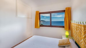 A bedroom with a bed, a window with curtains, a nightstand with a lamp, and a view of the sea. - Villetta Nuvola Azzurra (Trinità d'Agultu e Vignola)
