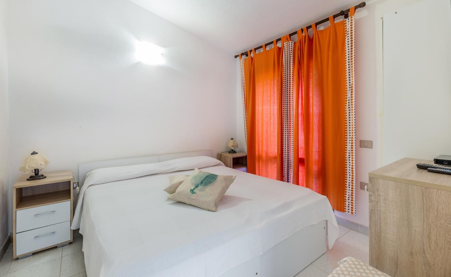 Et soveværelse med seng, natbord, lampe, skrivebord og et orange gardin.