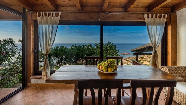 A wooden dining table with chairs, a bowl of fruit, and a view of the sea through large windows. - Villetta Hemingway (Trinità d'Agultu e Vignola)