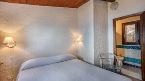 A bedroom with a bed, a chair, a wall-mounted lamp, and a bathroom visible through an open door. - Villetta Hemingway (Trinità d'Agultu e Vignola)