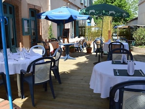 Eine Außenpatio mit Tischen und Stühlen, teilweise unter blauen und grünen Sonnenschirmen, mit einem Backsteinhaus im Hintergrund.