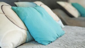 Una cama con múltiples almohadas en tonos blanco y verde azulado, un edredón gris con textura y un cabecero oscuro.