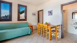 A room with a green sofa, wooden dining table with chairs, a small window, and a bathroom with a sink.