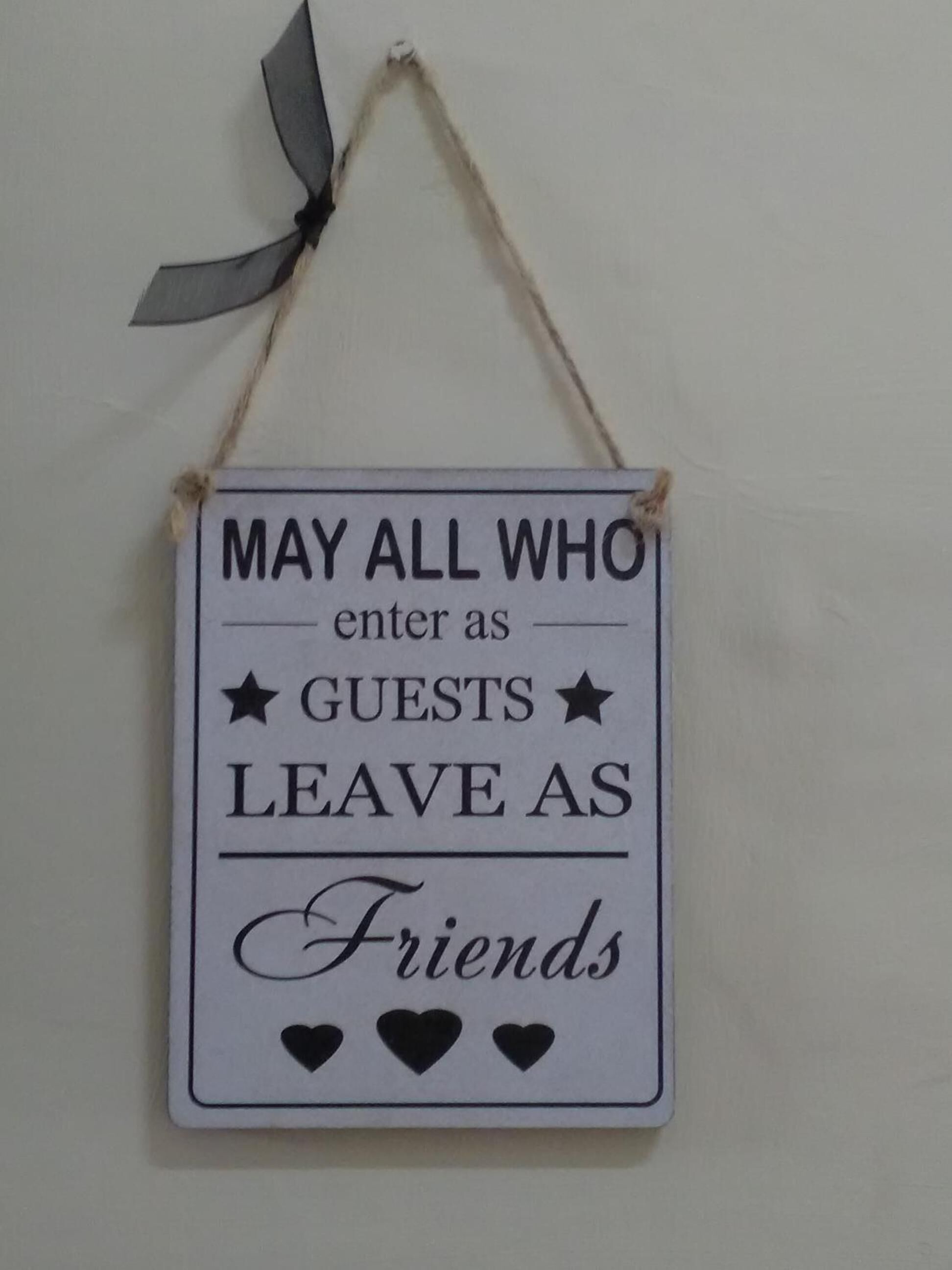 A sign with the text "MAY ALL WHO enter as GUESTS LEAVE AS Friends" and decorative hearts.