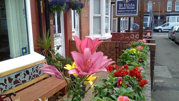 A bench with a tiled bench seat, a flower bed with pink lilies, yellow lilies, and red flowers, and a sign for St Hilda's Guest House. - St Hilda Guest House (Bridlington)