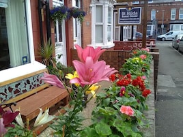 A bench with a tiled bench seat, a flower bed with pink lilies, yellow lilies, and red flowers, and a sign for St Hilda's Guest House.