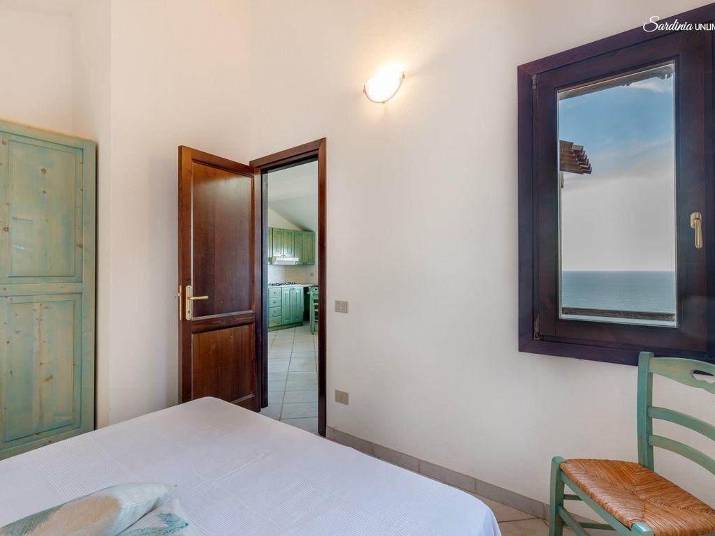 A bedroom with a bed, a wooden door, a green chair, and a window with a view of the sea.