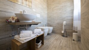 A modern bathroom with a large sink, toilet, and bidet. There is a wooden shelf with towels and a small basket. - Suite Simonetta (Sarzana)
