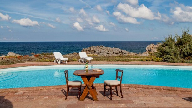 A poolside area with a table and chairs, overlooking the ocean. - Villa Mariù e Amelie (Aglientu)