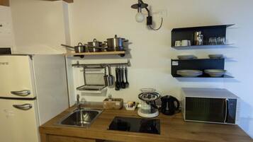 A compact kitchen with a white refrigerator, a microwave, and a sink. There are shelves above the counter holding dishes and utensils.