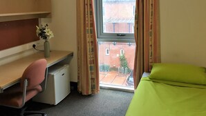 A small room with a desk, chair, and a bed. There is a window with curtains, a small fridge, and a plant on the desk. - Davies Court - Hostel (London)