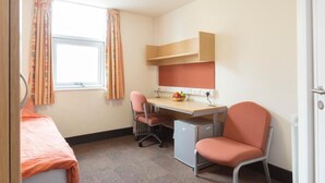 A small, well-lit room with a bed, a desk with a computer, a chair, and a small refrigerator. - Davies Court - Hostel (London)