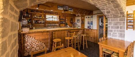 A rustic bar area with wooden furniture, stone walls, and a variety of bottles on shelves.