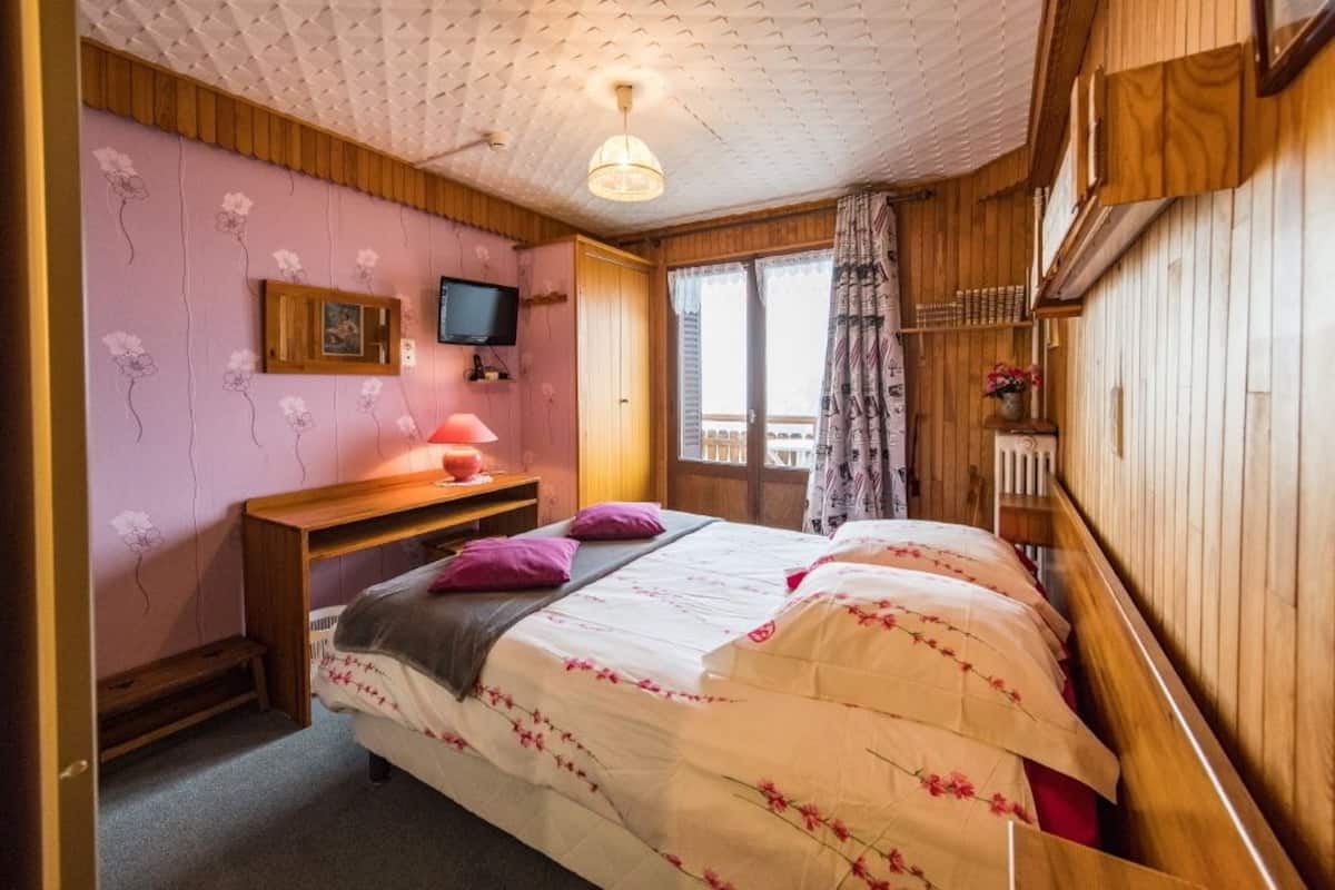 Une chambre à coucher avec un lit, un bureau, une télévision et une fenêtre avec des rideaux.