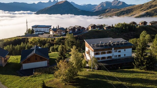 Un pueblo de montaña con casas de madera tradicionales, un edificio grande con techo inclinado y vistas a las montañas circundantes y nubes.