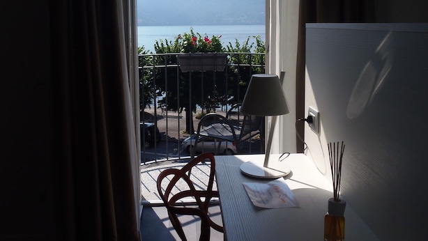 A room with a table, a lamp, and a bottle of reed diffuser. There is a view of a balcony with chairs and a table outside. - Albergo Dongo (Dongo)