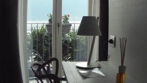 A room with a table, chair, lamp, and a vase with reed sticks. A window with curtains is visible, and there is a balcony with plants and a railing. - Albergo Dongo (Dongo)
