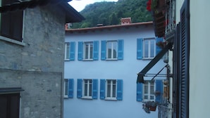 A building with multiple windows and blue shutters, a stone facade, and a balcony with potted plants. - Albergo Dongo (Dongo)