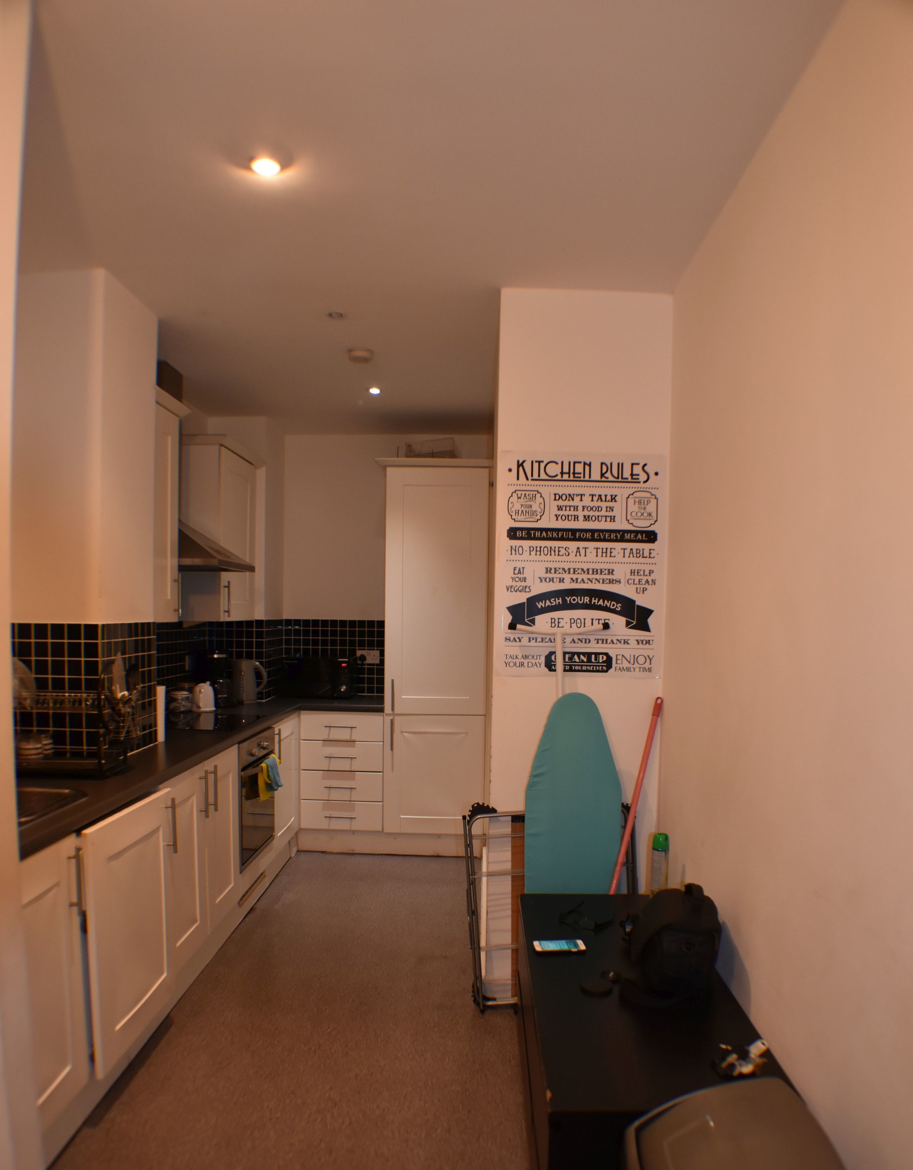 Una cocina compacta con armarios blancos, una encimera negra y un cartel en la pared con las normas de la cocina.
