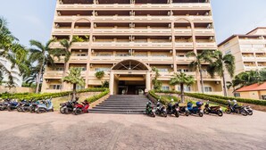 Et hotel med flere etager og en central indgang, omgivet af parkerede scootere og palmer.