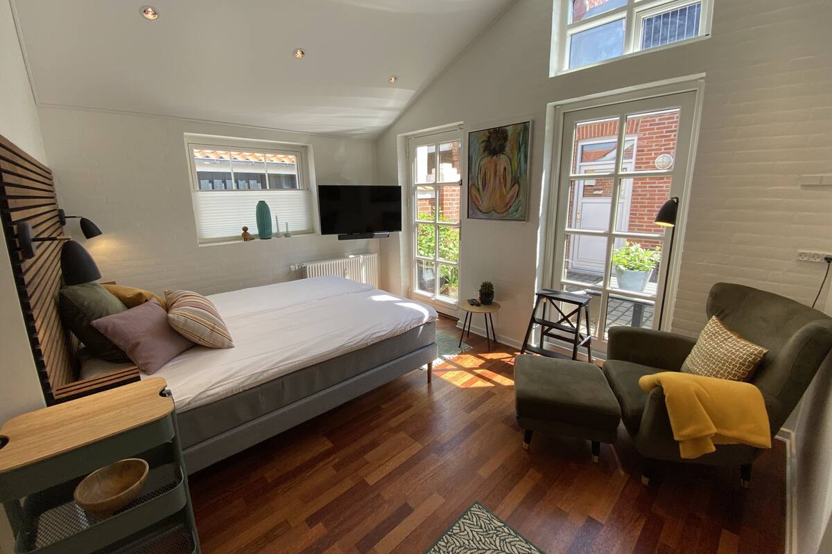A modern bedroom with a large bed, a sofa, and a TV. There are windows and a view of the outside.
